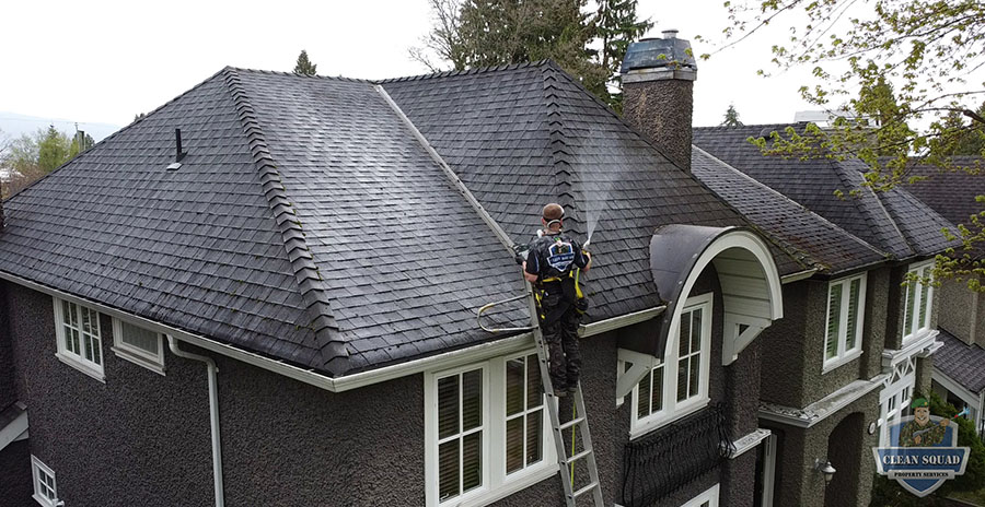 a drone photo of a clean squad employee spraying soft wash solution onto a shingle roof from a ladder